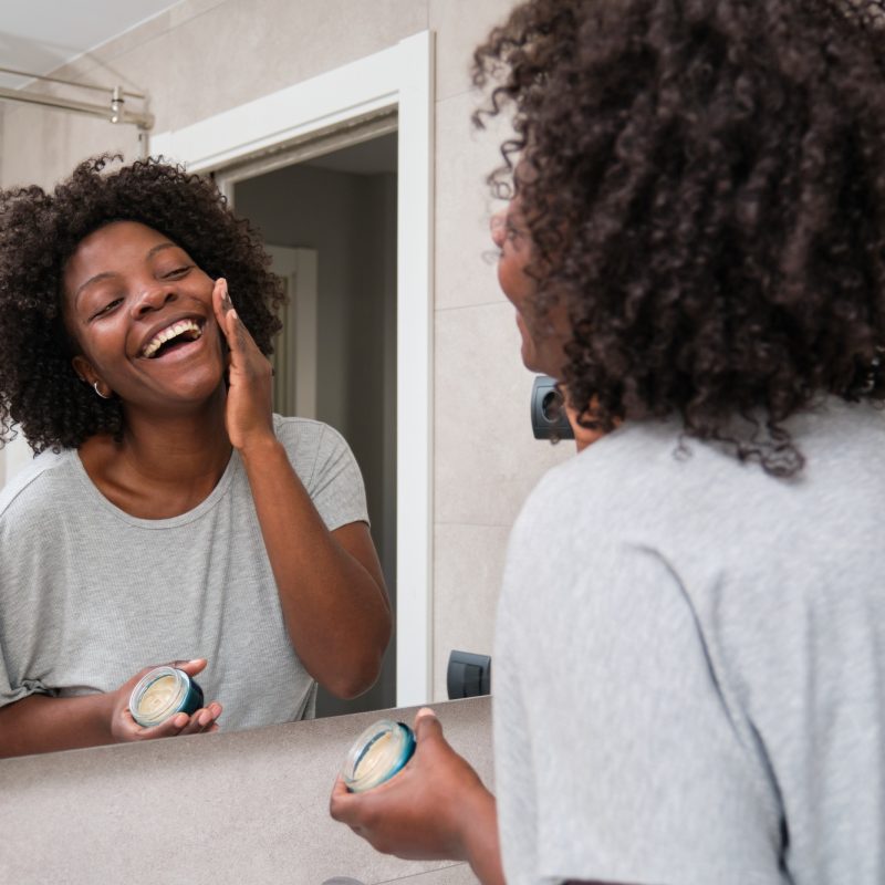 Smiling african woman applies eco-friendly facial cream in bathroom.